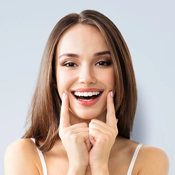A woman with a radiant smile, touching her teeth with her hand, against a light blue background.