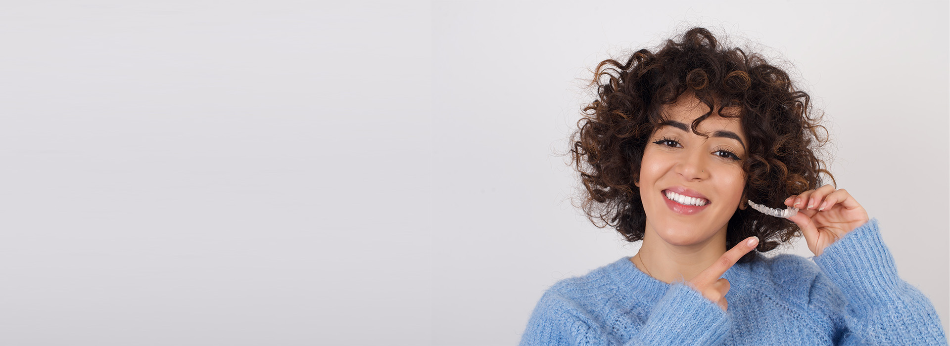 A woman with curly hair smiling at the camera, wearing a blue sweater, against a white background.