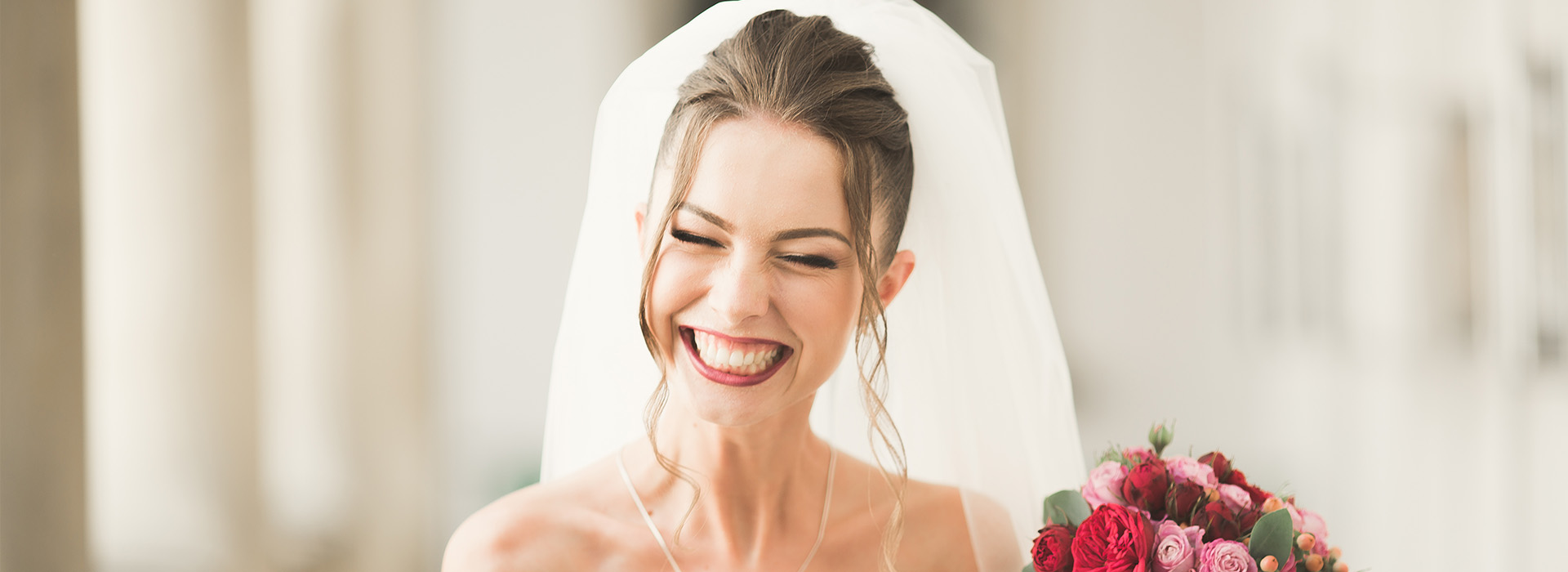 The image features a woman wearing a white wedding dress and veil, smiling and looking upwards with her mouth open, standing next to a man who appears to be her husband, both in front of a white pillar  behind them is a blurred background that suggests an indoor setting, likely a church or chapel.