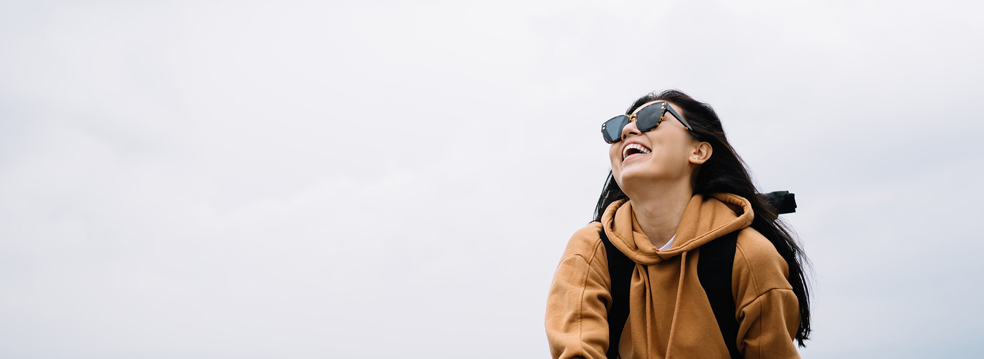The image shows a person standing outdoors with sunglasses on, wearing a backpack, smiling at the camera, and looking into the distance against a cloudy sky background.