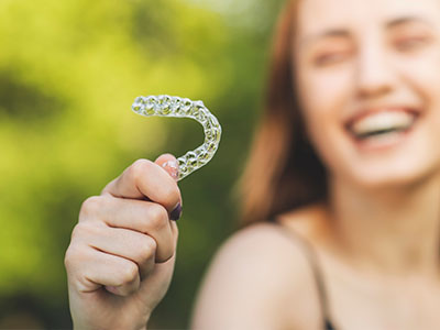 The image features a smiling young woman holding up a clear plastic dental retainer with a toothbrush-like bristle design, set against a blurred outdoor background.