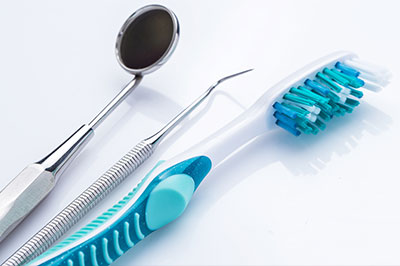 The image displays dental tools including toothbrushes with blue bristles, a metal dental mirror, and a pair of tweezers, all laid out on a white surface against a blurred background.