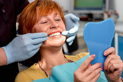 A woman receiving dental care, sitting in a dental chair with blue gloves on her hands holding a model tooth, while wearing a white gown.