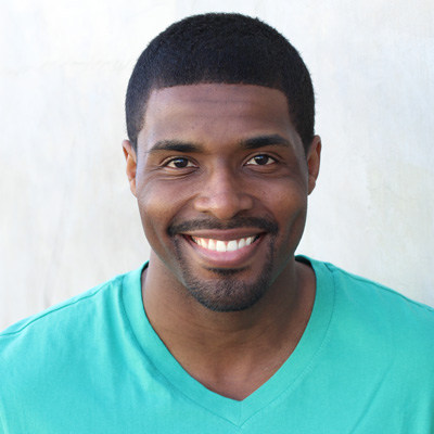 The image shows an older couple sitting closely together, smiling at each other, with the man wearing glasses and both dressed casually, set against a blurred outdoor background that suggests a warm climate.