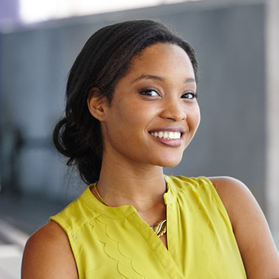 A young woman with a radiant smile, wearing a yellow blouse and posing against a backdrop of a building.