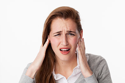 The image shows a woman with her hand on her head, possibly indicating pain or discomfort. She appears to be indoors, wearing a light-colored top and has a concerned expression.