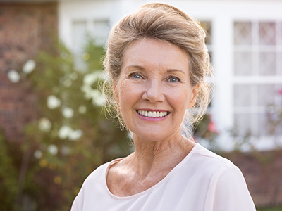 The image shows a woman standing outdoors in front of a house with white walls and a red brick chimney, smiling at the camera.