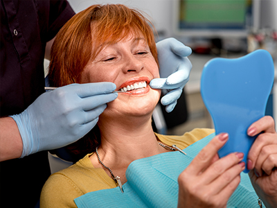 A woman seated in a dental chair, smiling at the camera while holding a blue mouth guard, with a dental hygienist performing a cleaning procedure.
