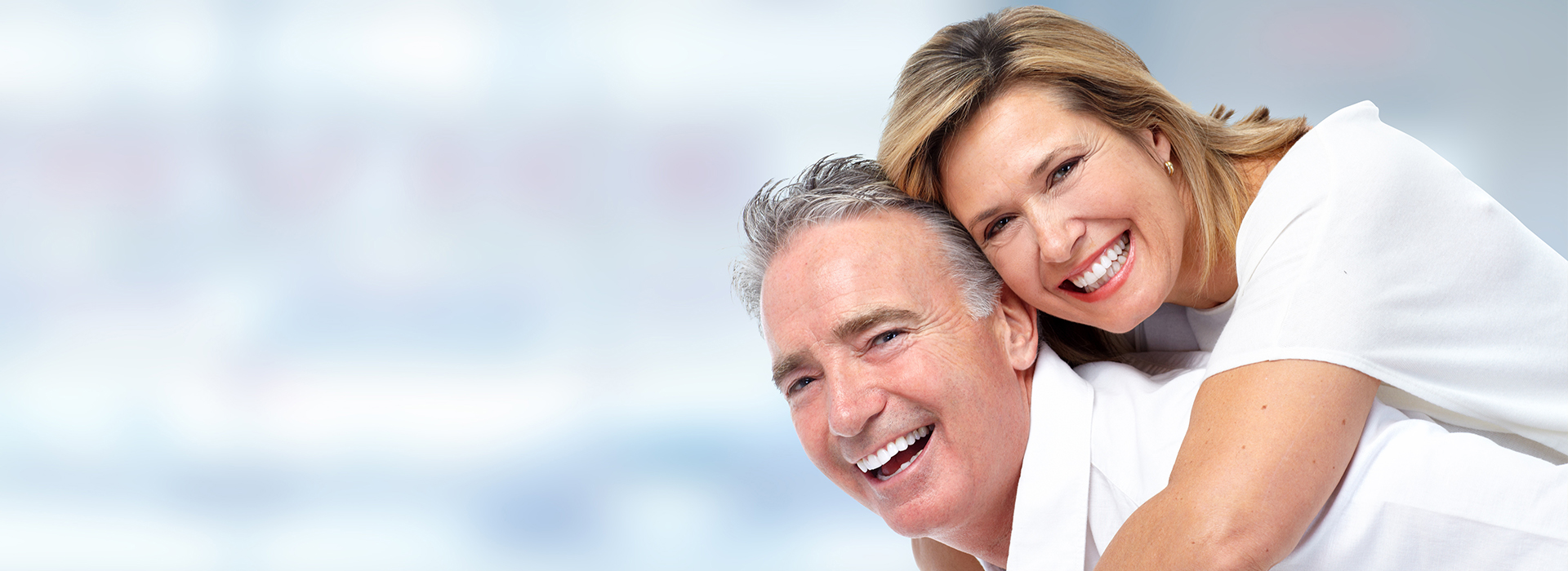 The image shows a man and a woman holding each other, both smiling, with a blurred background that suggests they are outdoors during daylight.