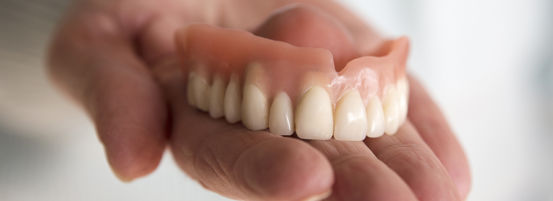 An adult human hand holding a set of artificial teeth with white enamel against a neutral background.