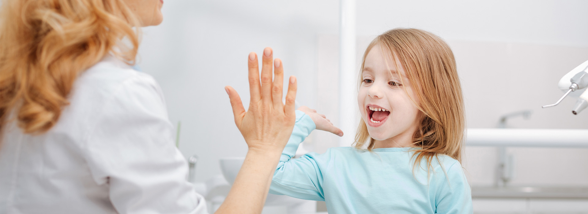 The image shows two individuals interacting with each other  one appears to be an adult woman wearing a white apron and smiling, while the other is a young girl who seems to be looking at her hand. They are in a room that resembles a dental office, as indicated by the presence of a sink and dental chair in the background.