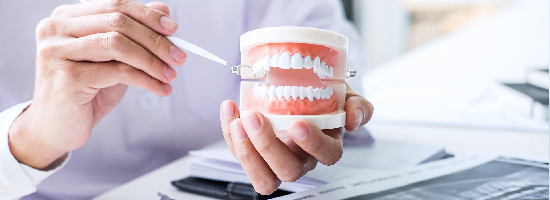 A person s hand holding a toothbrush with toothpaste on a desk while another hand holds a cup with a red substance, possibly dental floss, against a blurred background of a workspace.