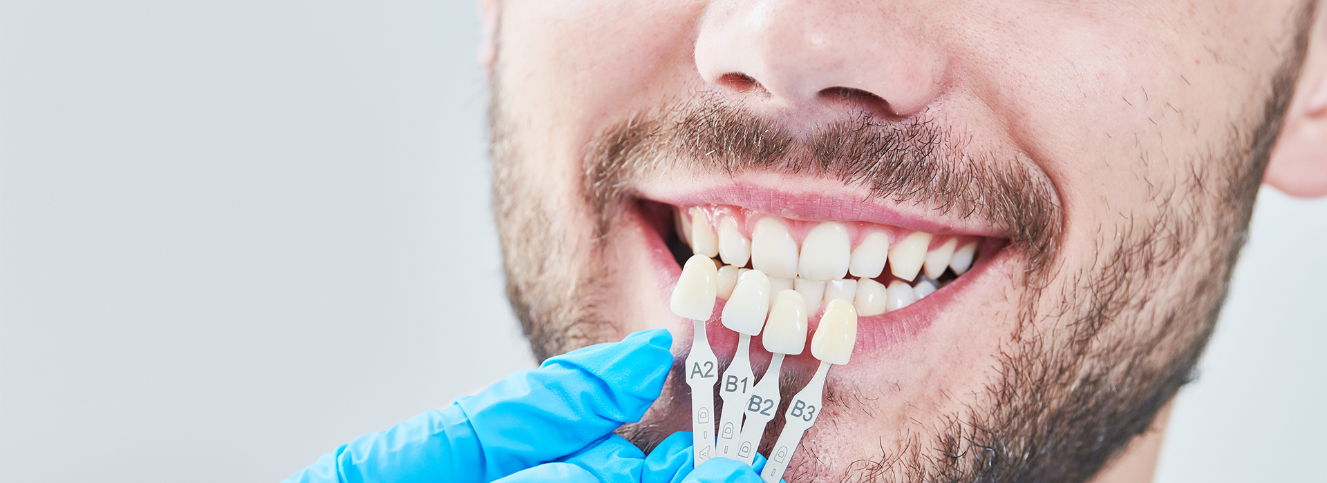 A man with a toothy smile, wearing a blue surgical mask, has his teeth checked by a dental professional using a magnifying glass.