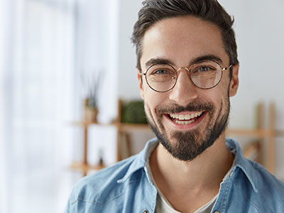 A man with a beard and glasses is smiling at the camera, wearing a blue shirt and standing against a white wall.