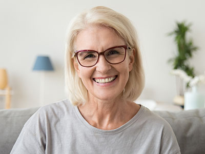 The image shows an older woman with short blonde hair smiling at the camera. She wears glasses and a light blue top, seated indoors with a neutral-toned background that includes a lamp and a plant.