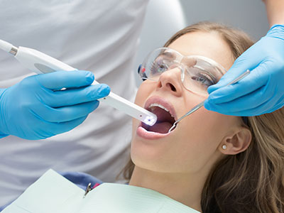 A dental hygienist is performing a teeth cleaning procedure on a woman s mouth using an electric toothbrush, with both parties wearing protective gloves and face masks.