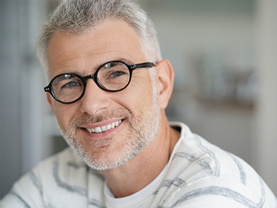 A man with glasses and gray hair is smiling at the camera, wearing a white t-shirt and looking directly ahead.