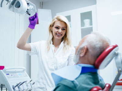 The image shows a dental professional assisting an elderly patient in a dental chair with medical equipment around them.