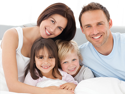 An alt text description of the image   A family of four, including two adults and two children, smiling together on a bed.