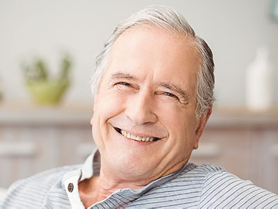 The image shows an elderly man with gray hair, smiling broadly, wearing a blue shirt, seated comfortably in a chair, with his eyes closed and hands resting on his chest.