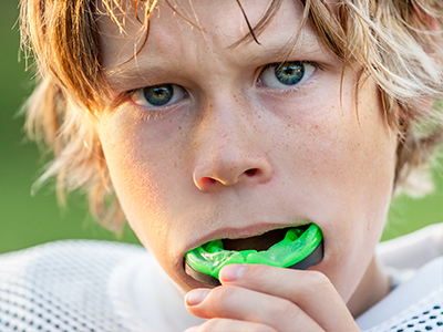 The image shows a young boy with blonde hair, wearing a football uniform, holding a green object near his mouth.