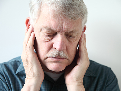 The image shows an older man with his eyes closed, holding his hand to his face, possibly in pain or discomfort, against a plain background.